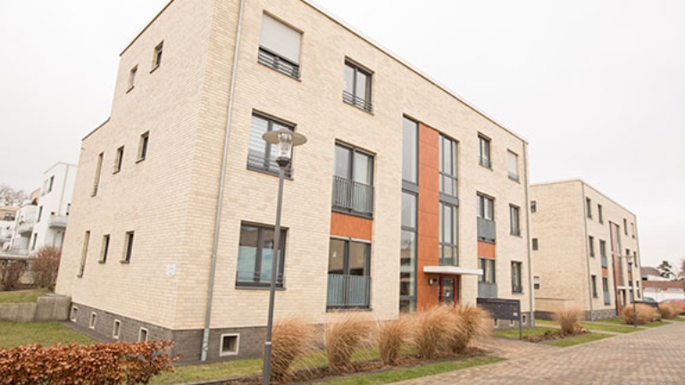 Zwei Gebäude eines Wohnquartiers mit heller Fassade, davor eine ruhige Straße und Vorgärten