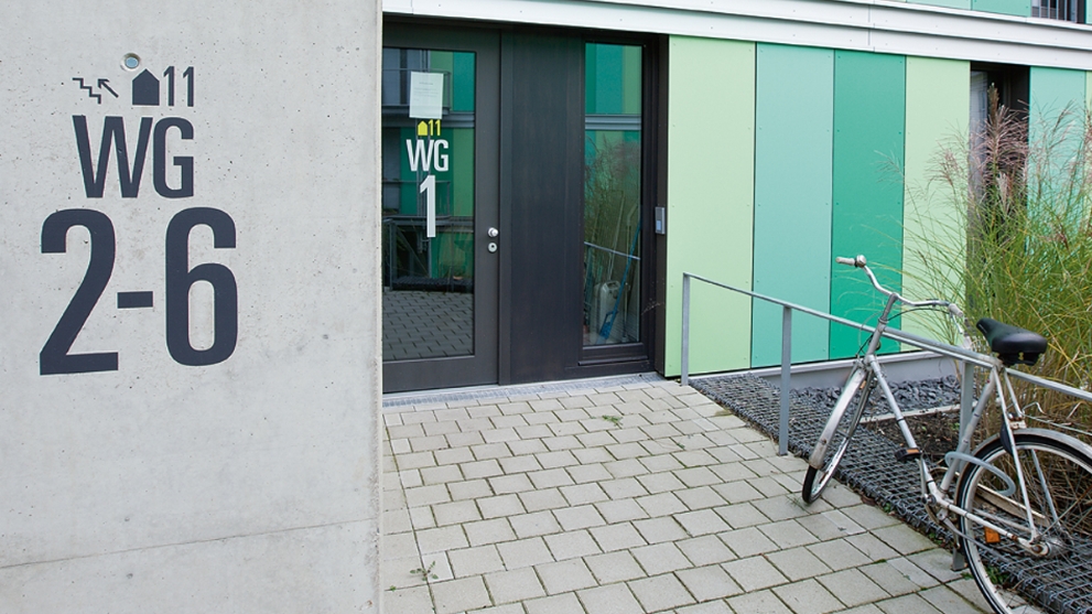 Grüne Fassade und Eingangsbereich eines Studentenwohnheims, davor ein gepflasterter Weg und ein Fahrrad