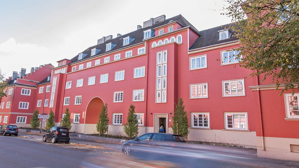 Sanierter Altbau-Gebäudekomplex mit strahlend roter Fassade, davor eine Straße, parkende Autos und einige Bäume