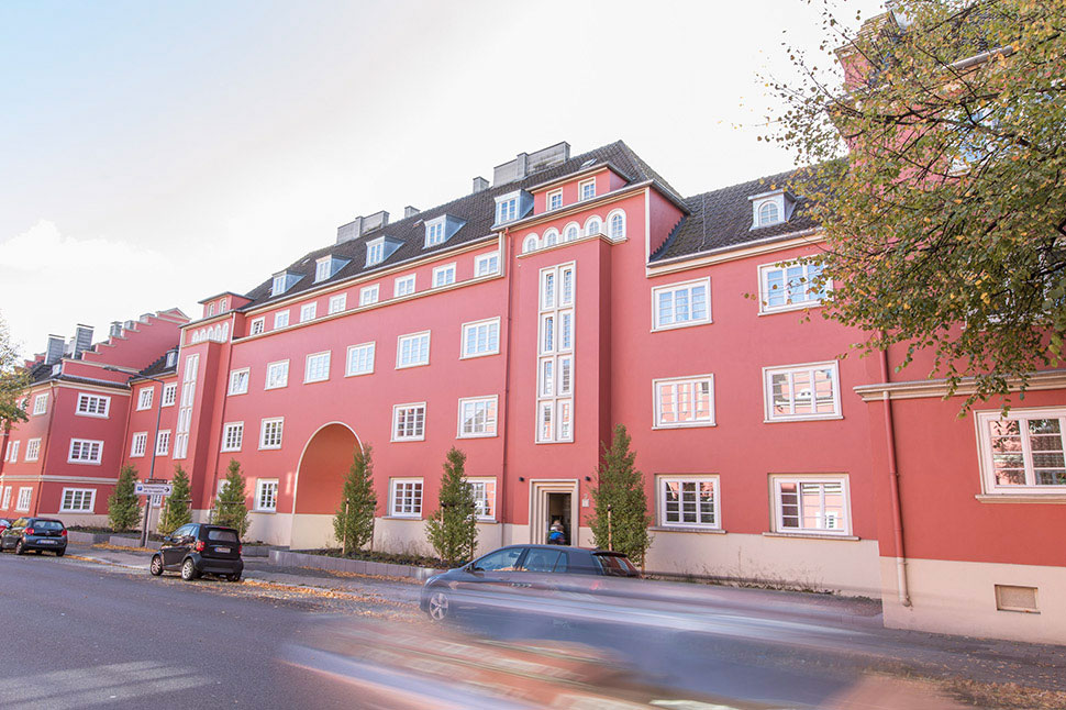 Sanierter Altbau-Gebäudekomplex mit strahlend roter Fassade, davor eine Straße, parkende Autos und einige Bäume