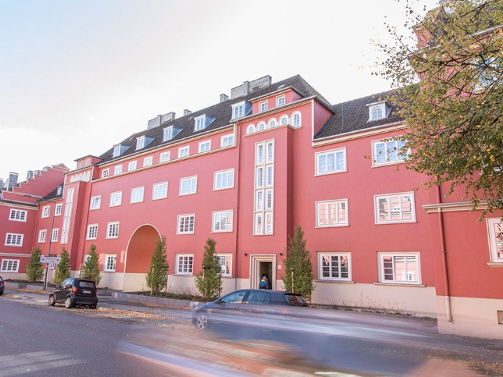 Sanierter Altbau-Gebäudekomplex mit strahlend roter Fassade, davor eine Straße, parkende Autos und einige Bäume
