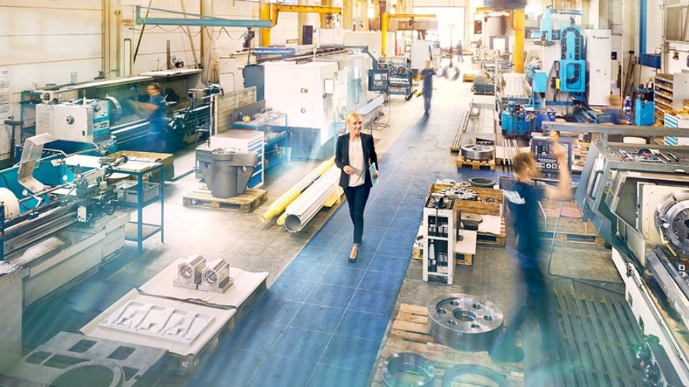 Unternehmerin Melanie Baum geht durch ihre Werkshalle. Metallteile und Maschinen sowie mehrere Mitarbeiter sind zu sehen.
