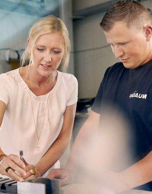 Unternehmerin Melanie Baum und ein Mitarbeiter in einer Werkstatt, sie zeigt ihm die Bearbeitung eines Metallteils.