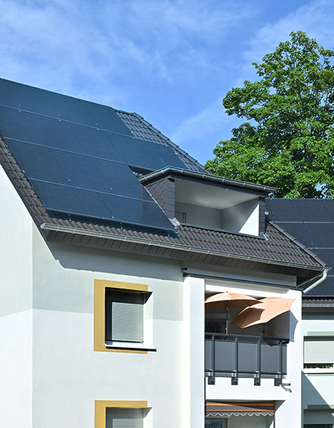 Modernes Wohnhaus mit großen Photovoltaikmodulen auf dem Dach, weißen Fassaden und Balkon; umgeben von grünen Bäumen an einem sonnigen Tag.