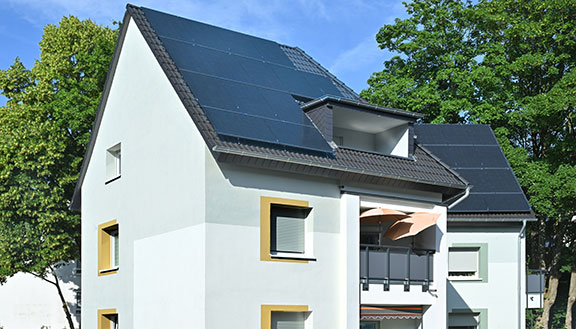 Modernes Wohnhaus mit großen Photovoltaikmodulen auf dem Dach, weißen Fassaden und Balkon; umgeben von grünen Bäumen an einem sonnigen Tag.
