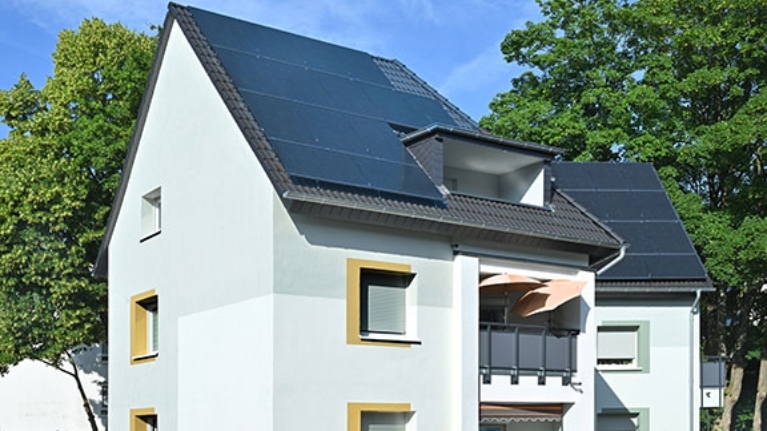 Modernes Wohnhaus mit großen Photovoltaikmodulen auf dem Dach, weißen Fassaden und Balkon; umgeben von grünen Bäumen an einem sonnigen Tag.