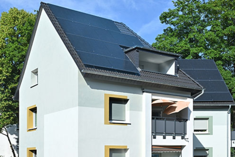 Modernes Wohnhaus mit großen Photovoltaikmodulen auf dem Dach, weißen Fassaden und Balkon; umgeben von grünen Bäumen an einem sonnigen Tag.