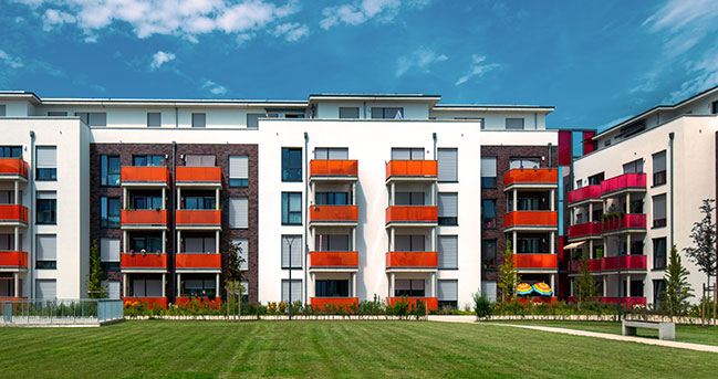 Moderne Wohnanlage mit weißen und dunkel verklinkerten Fassaden, farbigen Balkonen und großer Grünfläche davor unter blauem Himmel mit Wolken.