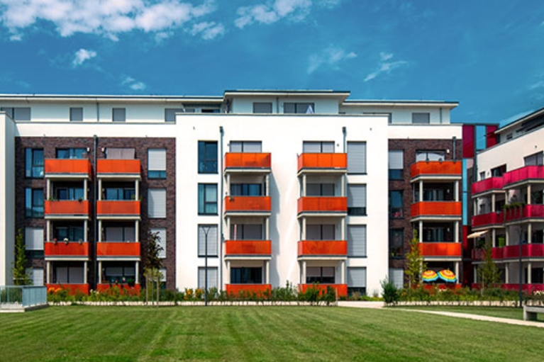 Moderne Wohnanlage mit weißen und dunkel verklinkerten Fassaden, farbigen Balkonen und großer Grünfläche davor unter blauem Himmel mit Wolken.