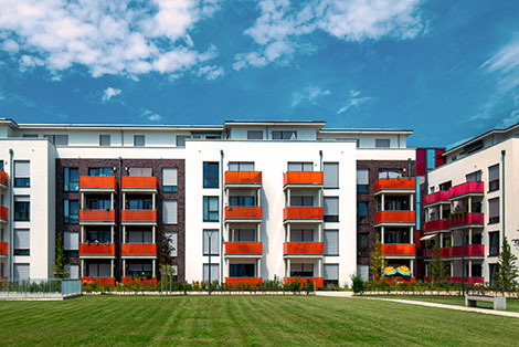 Moderne Wohnanlage mit weißen und dunkel verklinkerten Fassaden, farbigen Balkonen und großer Grünfläche davor unter blauem Himmel mit Wolken.