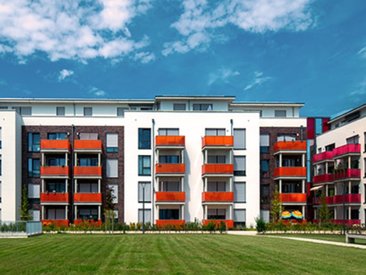 Moderne Wohnanlage mit weißen und dunkel verklinkerten Fassaden, farbigen Balkonen und großer Grünfläche davor unter blauem Himmel mit Wolken.