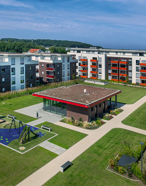 Weitläufige moderne Wohnanlage mit mehreren Mehrfamilienhäusern, grünem Innenhof, Spielplatz, Wegen und Gemeinschaftsgebäude unter blauem Himmel.