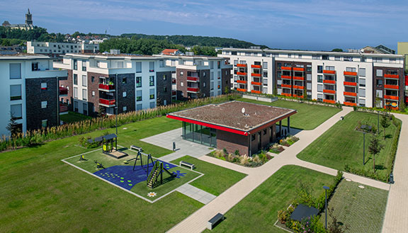 Weitläufige moderne Wohnanlage mit mehreren Mehrfamilienhäusern, grünem Innenhof, Spielplatz, Wegen und Gemeinschaftsgebäude unter blauem Himmel.