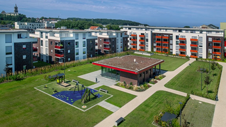 Weitläufige moderne Wohnanlage mit mehreren Mehrfamilienhäusern, grünem Innenhof, Spielplatz, Wegen und Gemeinschaftsgebäude unter blauem Himmel.