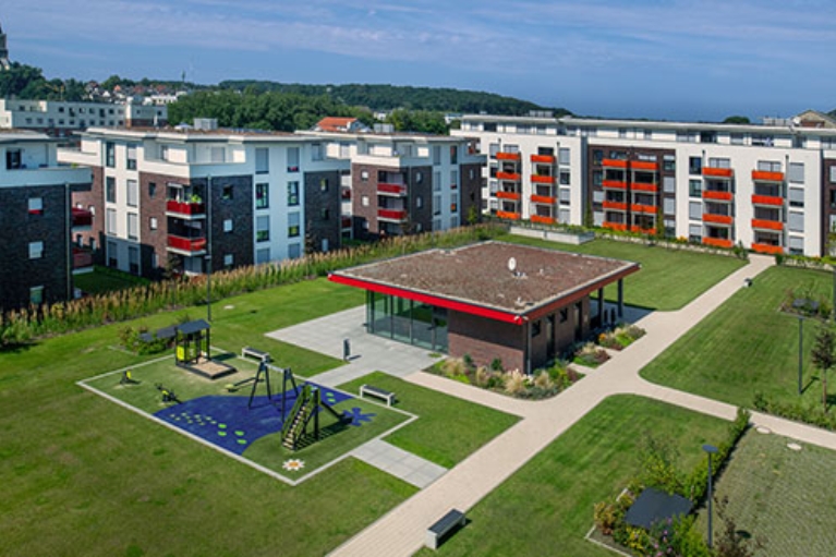 Weitläufige moderne Wohnanlage mit mehreren Mehrfamilienhäusern, grünem Innenhof, Spielplatz, Wegen und Gemeinschaftsgebäude unter blauem Himmel.