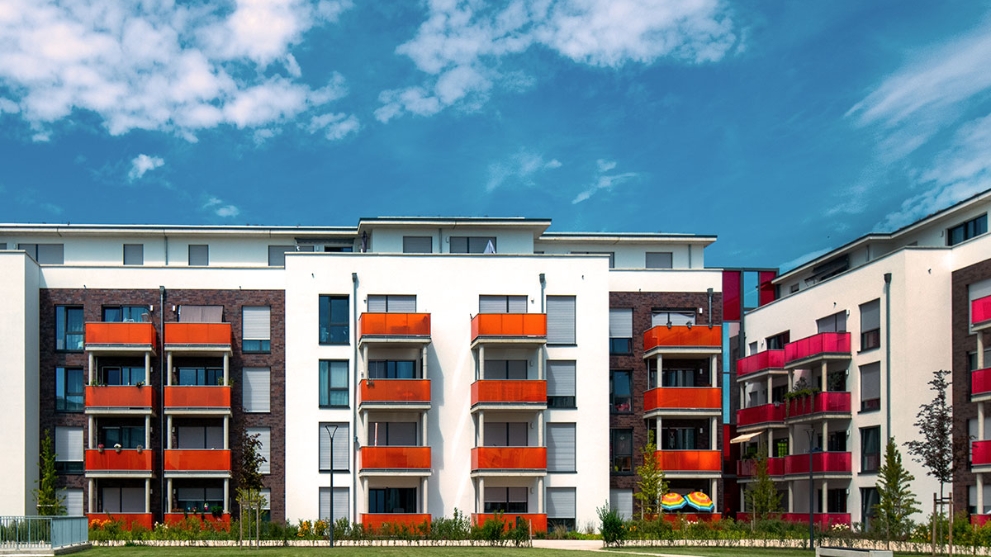 Moderne Wohnanlage mit weißen und dunkel verklinkerten Fassaden, farbigen Balkonen und großer Grünfläche davor unter blauem Himmel mit Wolken.