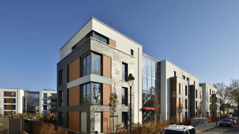 Modernes, neu gebautes Wohnquartier, davor Vorgärten mit kleinen Bäumen, im Hintergrund ein Spielplatz