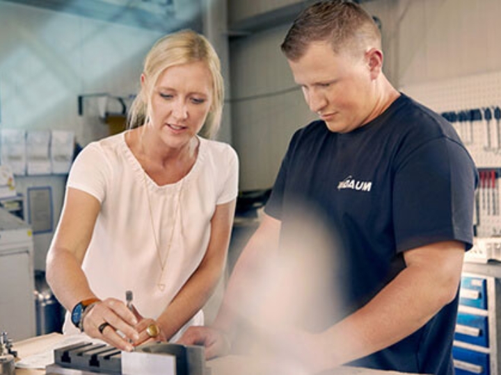 Unternehmerin Melanie Baum und ein Mitarbeiter in einer Werkstatt, sie zeigt ihm die Bearbeitung eines Metallteils.