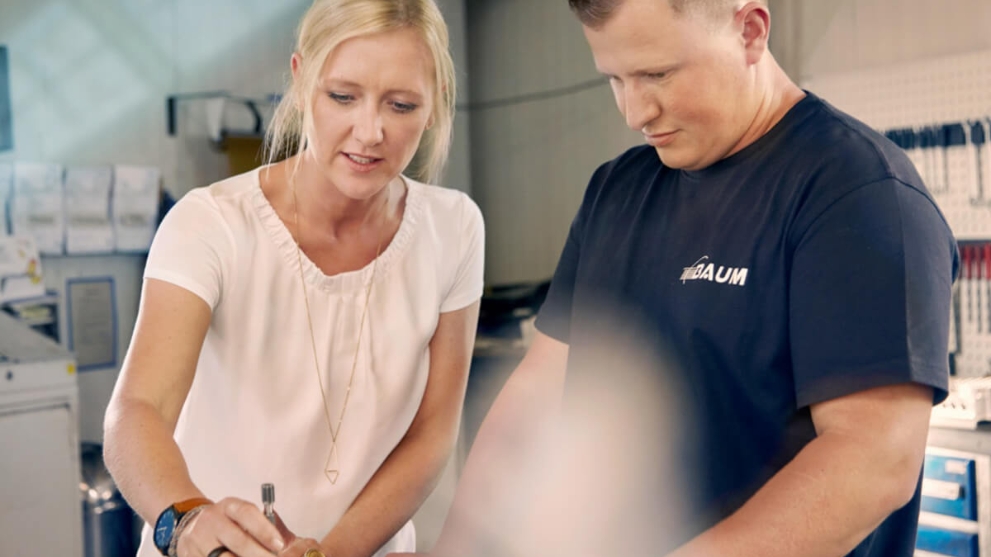 Unternehmerin Melanie Baum und ein Mitarbeiter in einer Werkstatt, sie zeigt ihm die Bearbeitung eines Metallteils.