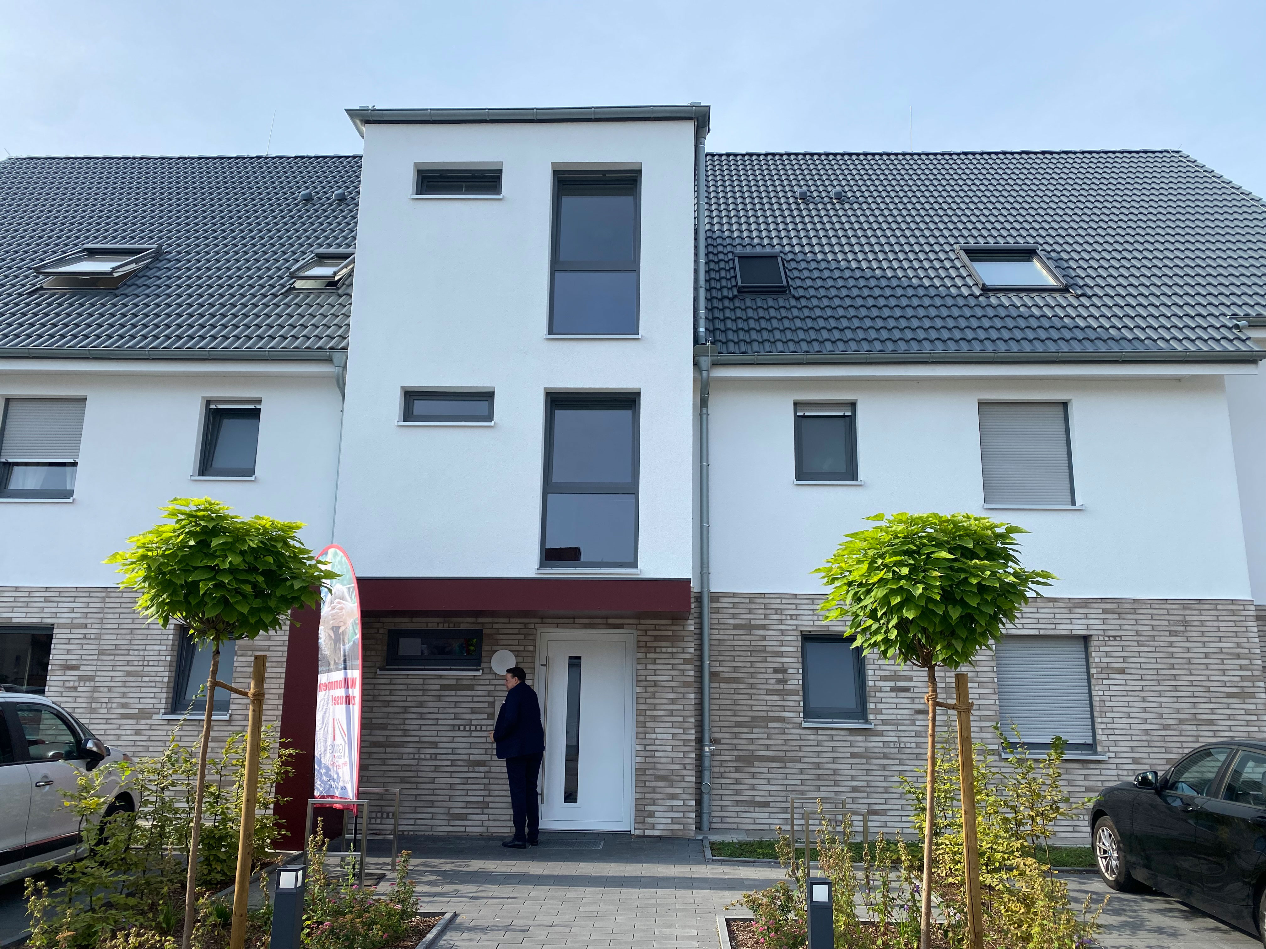 Neues Mehrfamilienhaus mit grau-weißer Fassade, davor parkende Autos und zwei kleine Bäume