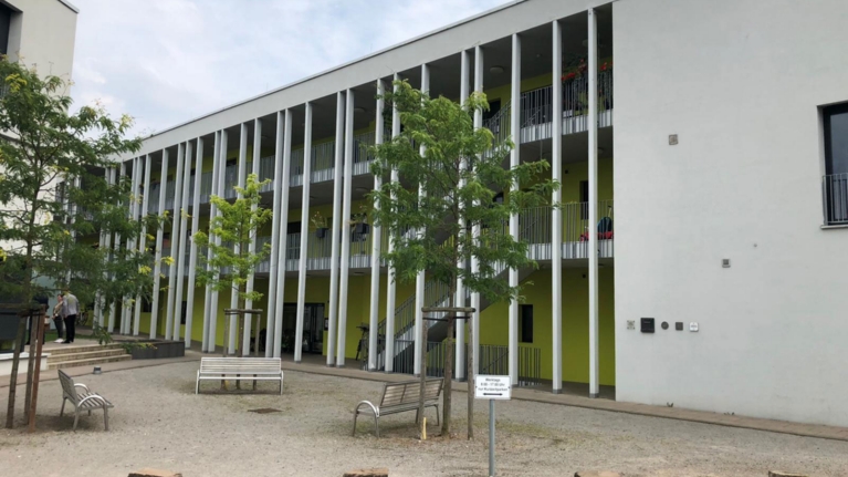 Rückseite eines Häuserblocks mit Säulen, davor ein Innenhof mit Bänken und Bäumen