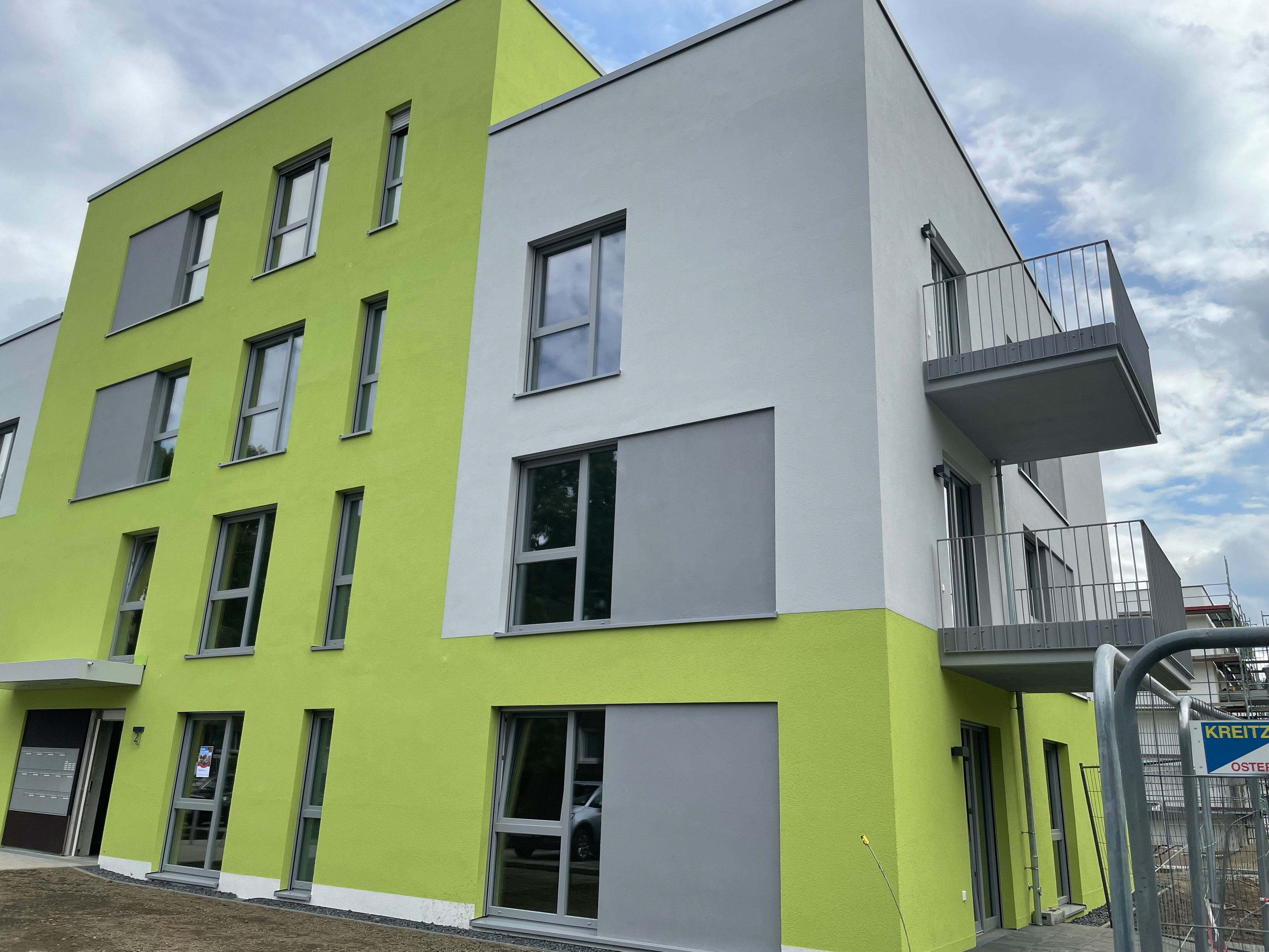 Neubau eines grün-grauen Wohnblocks mit Balkonen, vorne rechts ein Bauzaun