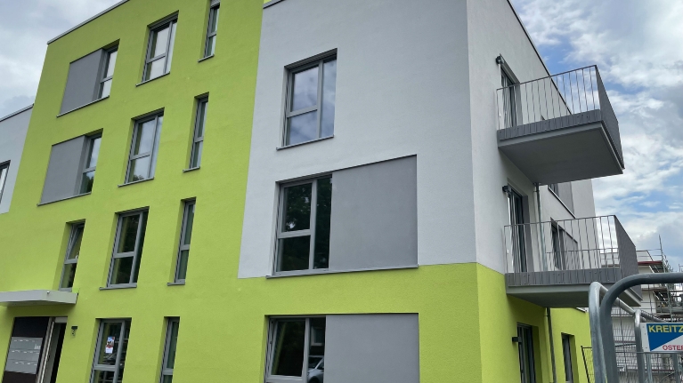 Neubau eines grün-grauen Wohnblocks mit Balkonen, vorne rechts ein Bauzaun