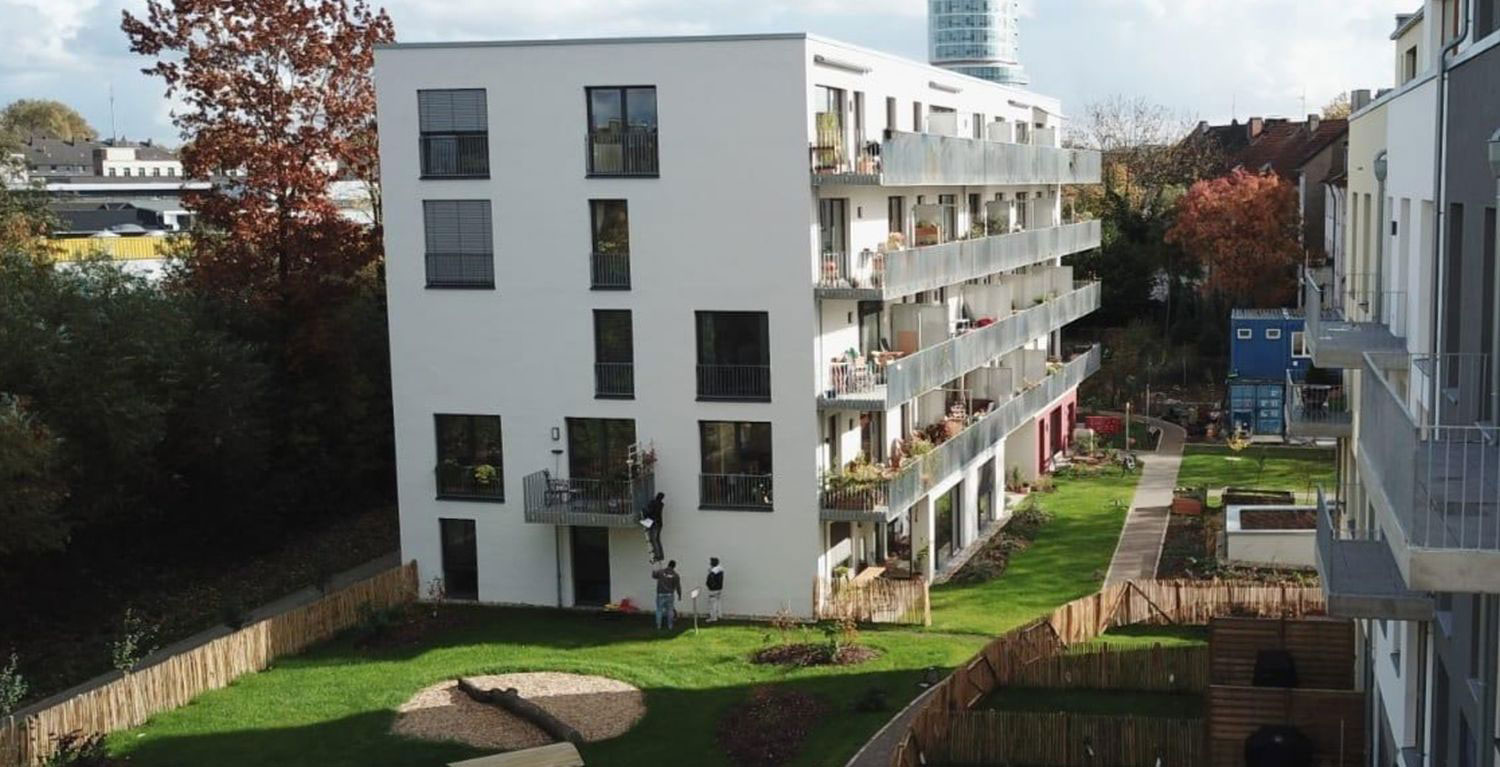 Vierstöckiger Wohnblock mit umliegendem Garten, im Hintergrund Bäume, Sträucher und ein Hochhaus