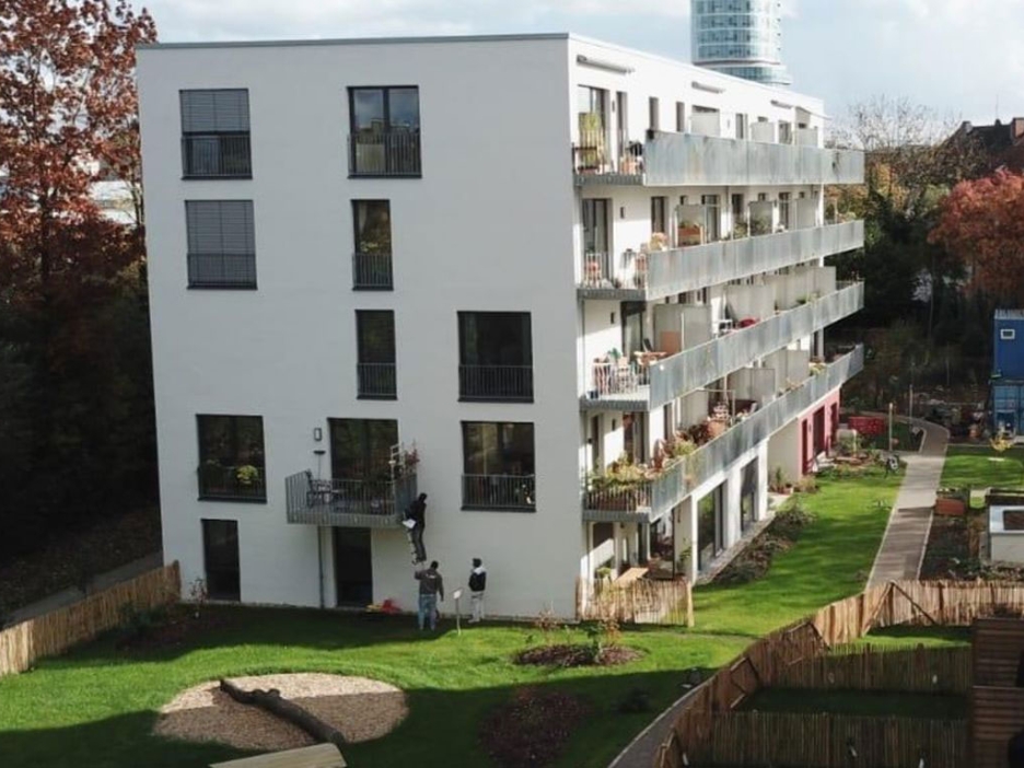 Vierstöckiger Wohnblock mit umliegendem Garten, im Hintergrund Bäume, Sträucher und ein Hochhaus
