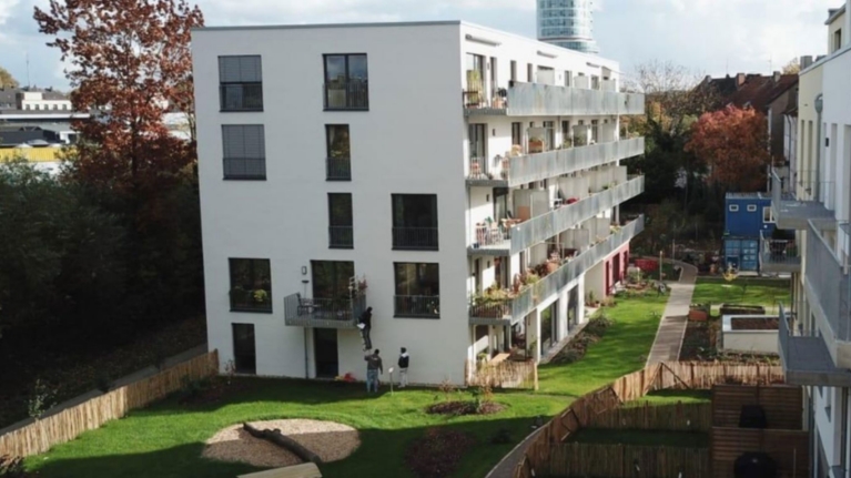 Vierstöckiger Wohnblock mit umliegendem Garten, im Hintergrund Bäume, Sträucher und ein Hochhaus