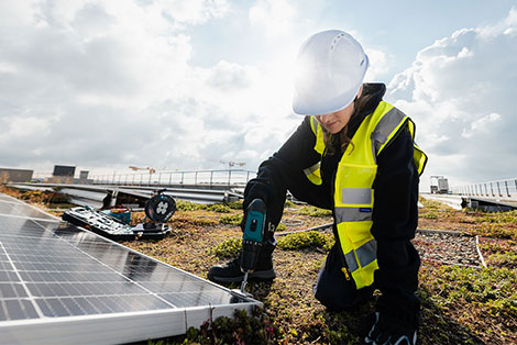 Ein Frau mit Schutzhelm und gelber Warnweste installiert auf einem Dach eine Solaranlage.