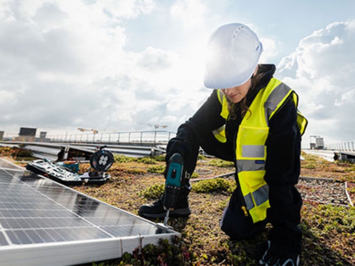 Ein Frau mit Schutzhelm und gelber Warnweste installiert auf einem Dach eine Solaranlage.