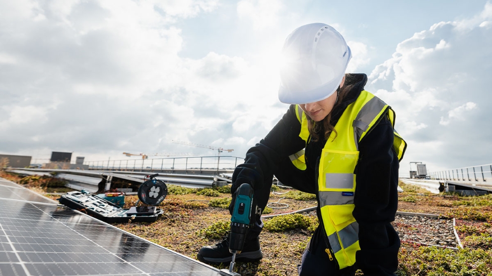 Ein Frau mit Schutzhelm und gelber Warnweste installiert auf einem Dach eine Solaranlage.