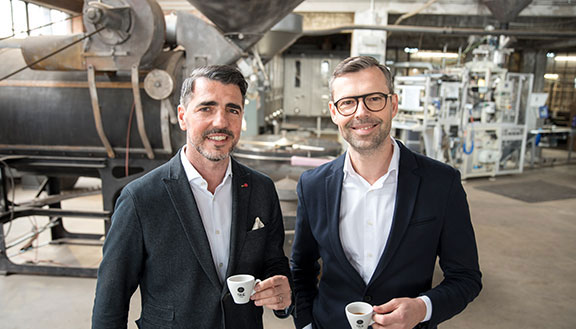 Die beiden Geschäftsführer von True Coffee lächeln mit einem Espresso in der Hand in die Kamera.