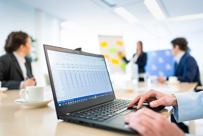 Vier Personen bei einer Präsentation in einem Besprechungsraum. Im Vordergrund ein Laptop mit einer Tabelle