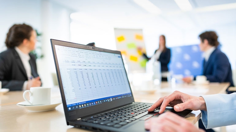 Vier Personen bei einer Präsentation in einem Besprechungsraum. Im Vordergrund ein Laptop mit einer Tabelle