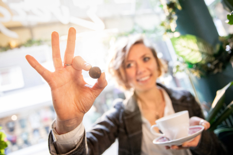 Eine Frau hält in der einen Hand eine Kaffeetasse, in der anderen einen kleinen kugelförmigen Snack. Sie lächelt dabei.