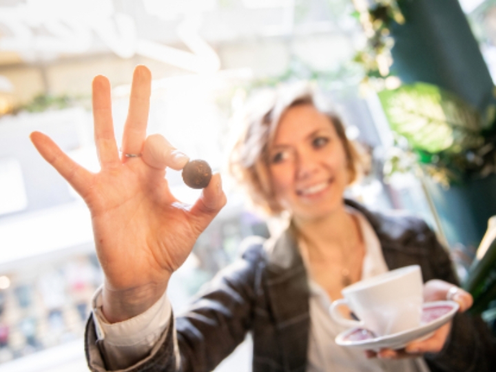 Eine Frau hält in der einen Hand eine Kaffeetasse, in der anderen einen kleinen kugelförmigen Snack. Sie lächelt dabei.