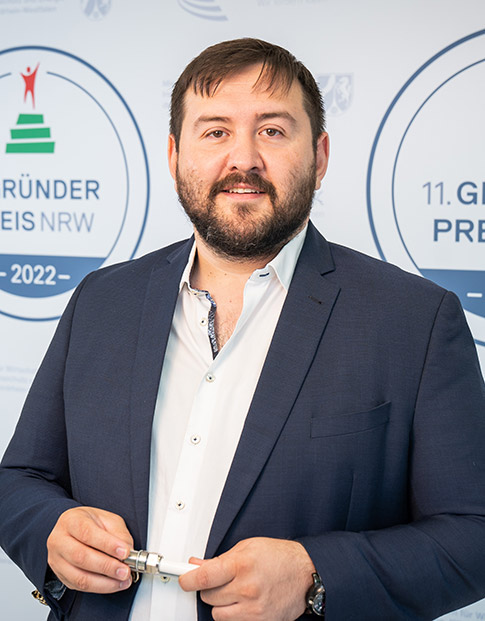 Der Nominierte des Gründerpreises 2022  Mesut Gökce der Onets GmbH steht vor der Gründerpreis Medienwand und hält ein Bauteil in seiner Hand.