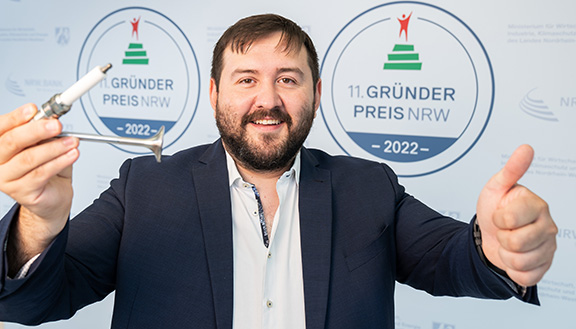 Der Nominierte des Gründerpreises 2022  Mesut Gökce der Onets GmbH steht vor der Gründerpreis Medienwand und hält ein Bauteil in seiner Hand.
