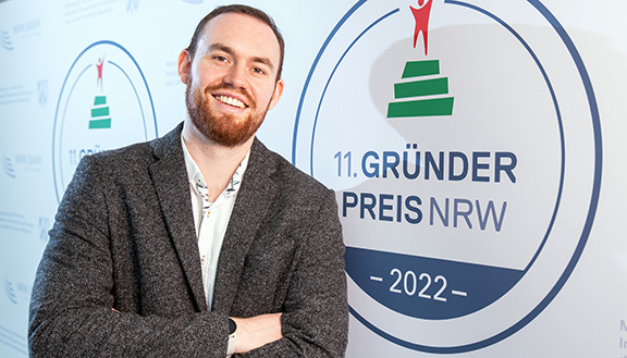 Der Nominierte des Gründerpreises 2022 Richard Haas der FibreCoat GmbH steht mit verschränkten Armen und einem Lächeln vor der Medienwand auf der das Gründerpreis-Logo zu sehen ist.