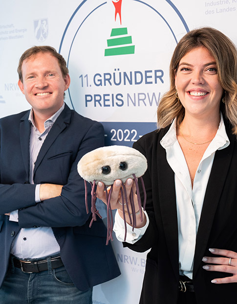 Die Nominierten des Gründerpreises 2022  Dr. Christian Schwarz und Teresa Harris der Numaferm GmbH halten ein Peptide- und Proteinmolekül-Stofftier in der Hand. Es hat lange Fäden und einem runden Kopf.