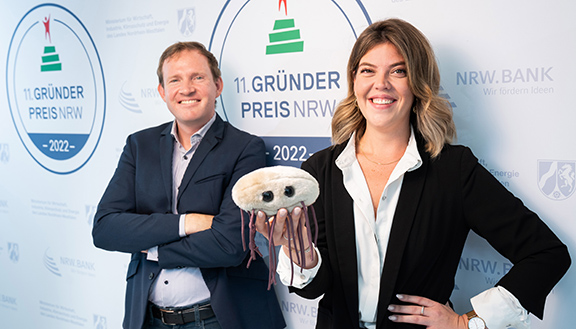 Die Nominierten des Gründerpreises 2022  Dr. Christian Schwarz und Teresa Harris der Numaferm GmbH halten ein Peptide- und Proteinmolekül-Stofftier in der Hand. Es hat lange Fäden und einem runden Kopf.