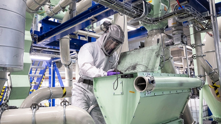 Eine Person in Schutzanzug und Helm arbeitet an einer großen Industrieanlage mit Rohren und Maschinen, beugt sich über einen geöffneten Behälter.