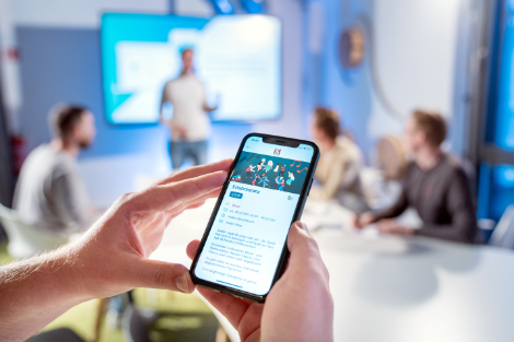 Im Hintergrund unscharf: Vier Menschen in einem Konferenzraum. Im Vordergrund hält jemand ein Smartphone in den Händen.