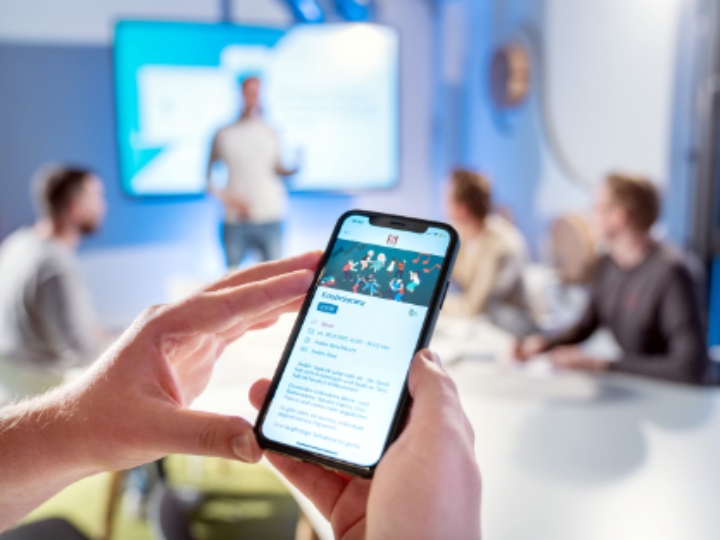 Im Hintergrund unscharf: Vier Menschen in einem Konferenzraum. Im Vordergrund hält jemand ein Smartphone in den Händen.