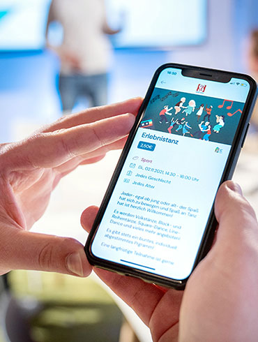 Das Foto zeigt Hände, die ein Smartphone halten, auf dem die Oberfläche der App Tremaze zu finden ist. Im Hintergrund sind vier Männer zu sehen, die ein Meeting abhalten.