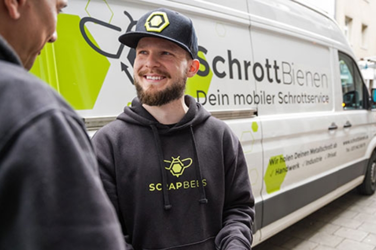 Mann im Scrapbees-Hoodie und Cap lächelt im Gespräch, im Hintergrund ein Transporter mit Firmenlogo.