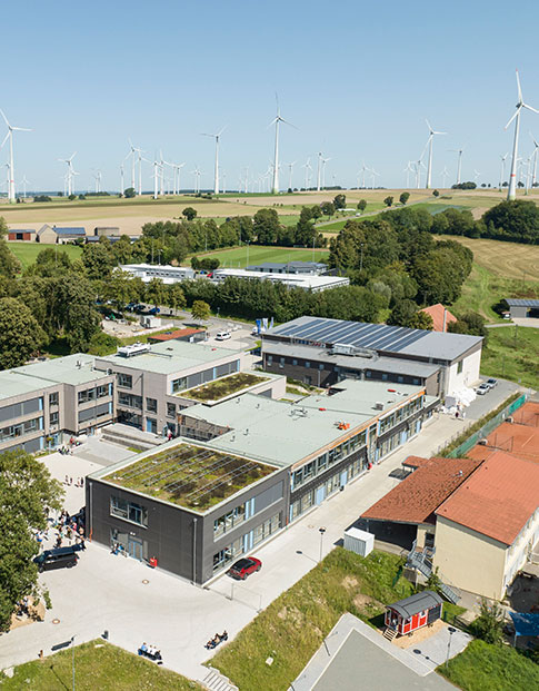 Moderne Campusgebäude mit begrünten Dächern und Windrädern im Hintergrund.