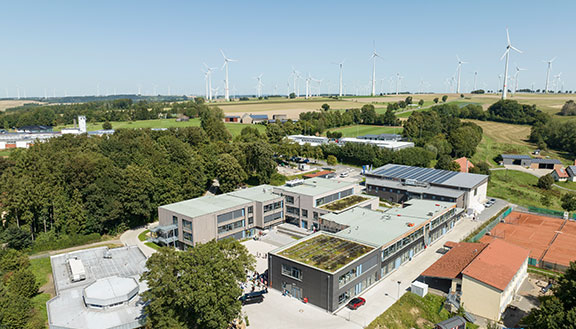 Moderne Campusgebäude mit begrünten Dächern und Windrädern im Hintergrund.